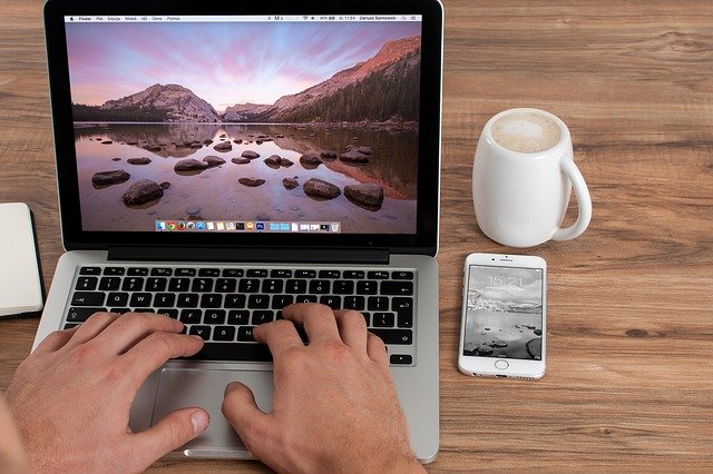 Laptop, Tasse Kaffee und Smartphone auf einem Schreibtisch; Hände, die die Tastatur des Laptops bedienen: Wenn Sie ein 80000 Euro Darlehen benötigen, können Sie heutzutage ganz leicht online einen günstigen 80000 Euro Kredit beantragen!