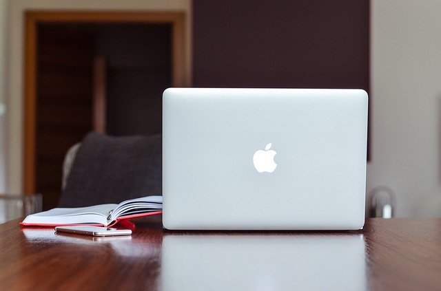 Macbook, rotes Notizbuch und Stift auf einem Schreibtisch; im Hintergrund ein Schreibtisch: Wenn Sie einen günstigen Privatkredit benötigen, dann schauen Sie sich doch mal den Bank of Scotland Ratenkredit genauer an!