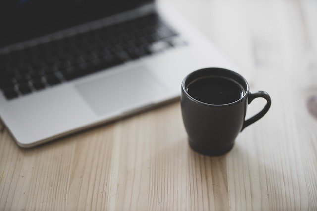 Schwarze Tasse mit Kaffee und Laptop auf einem Schreibtisch: Wenn Sie einen Kredit zur freien Verwendung benötigen, dann können Sie noch heute den flexiblen Degussa Bank Privatkredit online beantragen!