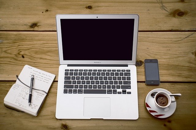 Notizblock mit Stift, Laptop, Smartphone und Tasse Kaffee auf einem Holztisch: Wenn Sie selbstständig sind und Ihr Geschäft ankurbeln möchten, kann Ihnen der günstige Postbank Business Kredit evtl. behilflich dabei sein!