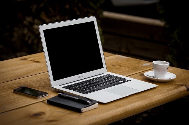 Laptop, Notizblock und Tasse Kaffee auf einem Holztisch: Wenn Sie Geld benötigen, können Sie den günstigen Postbank Kredit bequem online beantragen!